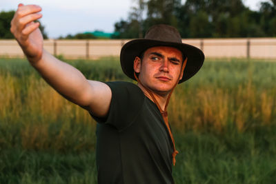 Young man serious in cowboy hat showing flying hands against green grass. male on sky background