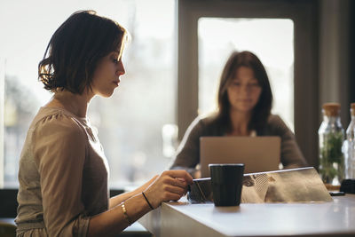 Side view of businesswoman reading document at desk with coworker in background