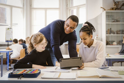 Smiling male teacher doing e-learning with female students in classroom