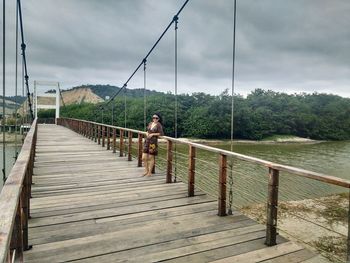 Young woman with hands on hip standing on footbridge against cloudy sky