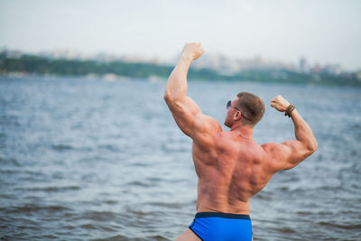 Rear view of shirtless bodybuilder flexing muscles while standing against sea