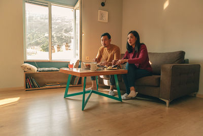 Full length of couple eating breakfast while watching tv sitting at home