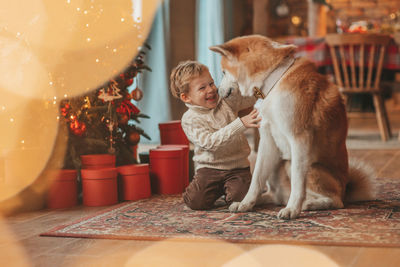 Candid authentic happy little boy in knitted beige sweater hugs dog with bow tie at home on xmas