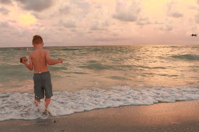 Boy standing on beach against sky during sunset