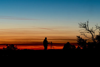 Silhouette photographer standing by trees against sky during sunset taking photo