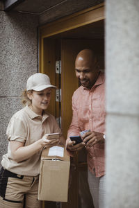 Young delivery woman and male customer using smart phones at doorway