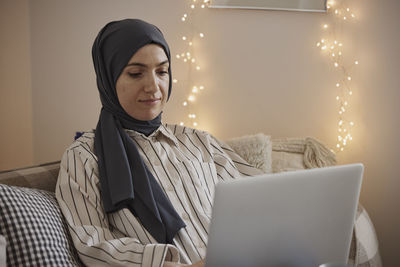 Young female freelancer wearing hijab and working on laptop in living room