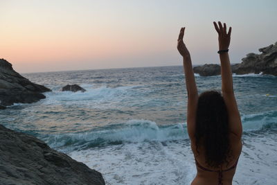 Rear view of woman with arms raised by sea against sky during sunset