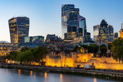 Illuminated buildings by river against sky in city at dusk