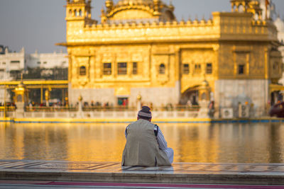 Rear view of man looking at golden temple while sitting on retaining wall by pond