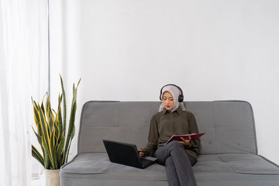 Portrait of young man using laptop while sitting against wall