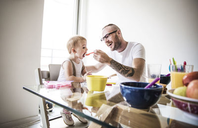 Happy father feeding daughter at breakfast table against brightly lit wall