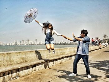 Man holding woman jumping on footpath by sea in city