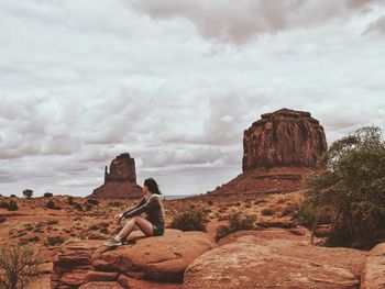 Side view of woman sitting on rock at landscape against cloudy sky