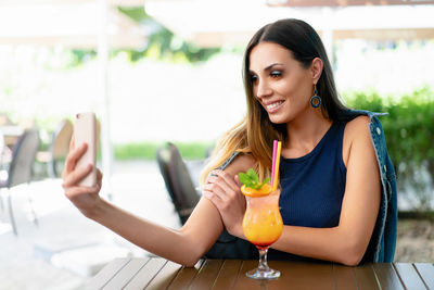 Young woman taking selfie through smart phone while sitting at sidewalk cafe