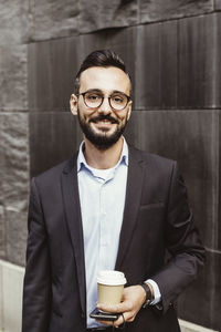 Portrait of confident businessman with disposable cup standing against wall