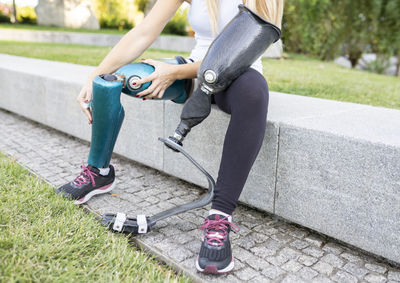 Unrecognizable crop female runner changing leg prosthesis for training while sitting on stone border in urban park