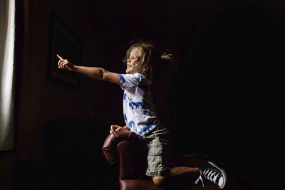 Smiling girl pointing towards window while kneeling on armchair in darkroom