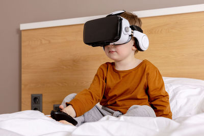 Little adorable boy sitting on bed at home with vr headset and playing interactive video game