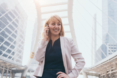 Smiling young woman using mobile phone while standing against wall