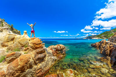 Rear view of woman standing on rock formations by sea against blue sky