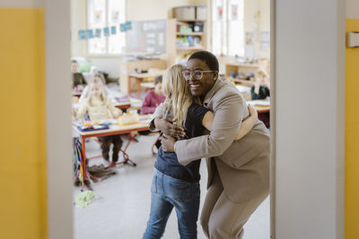 Smiling female teacher and schoolgirl embracing each other at doorway in classroom