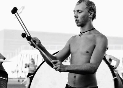 Shirtless young man holding drumsticks while standing against drum