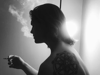 Side view of young tattooed man smoking cigarette in illuminated room