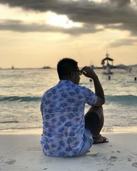 Rear view of man sitting at beach against sky during sunset