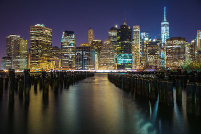 Wooden posts in sea against illuminated manhattan