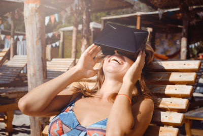 Cheerful young woman using virtual reality simulator while sitting on lounge chair at beach