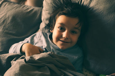 A little boy hides under the covers in bed and smiles.