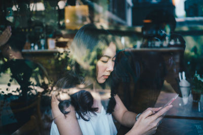 Side view of woman using mobile phone while sitting at table seen through glass window