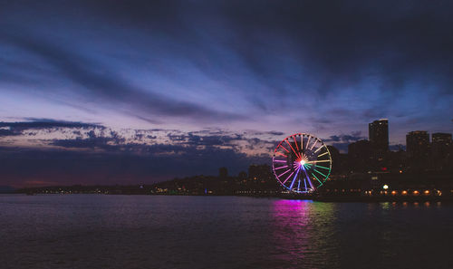 Illuminated ferris wheel by sea against sky at night