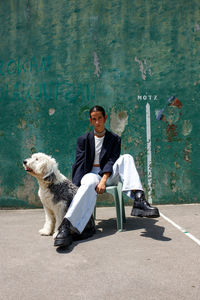 Full body of fashionable young ethnic female with dark hair in stylish outfit sitting on stool near adorable fluffy old english sheepdog near weathered wall on sunny day