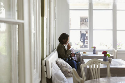 Side view of woman drinking coffee and using phone while resting at cafe