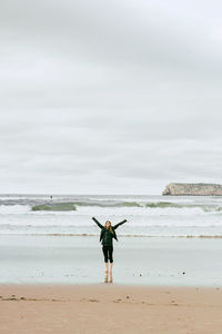 Full length of young woman with arms outstretched standing at beach against cloudy sky