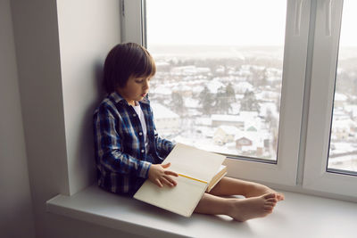 Boy child sits on the window of a house with a book in winter on a high floor