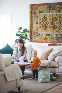 Female professional freelancing while daughter looking at papers on table in living room