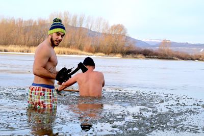 Shirtless men breaking ice in frozen lake against sky