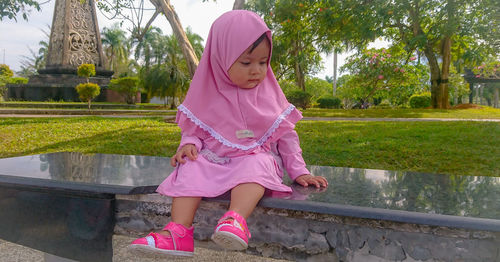 Full length of cute baby girl wearing hijab sitting on retaining wall