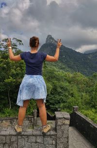 Woman gesturing peace sign while standing on retaining wall against mountains