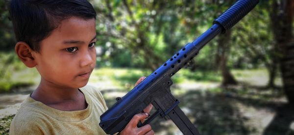 Boy looking away while holding toy gun against trees