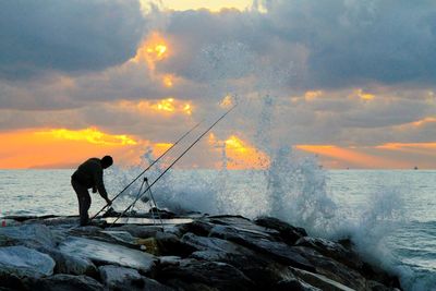 Man fishing in sea against sky during sunset