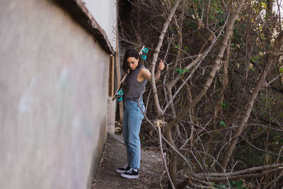 Full length of young woman holding skateboard while standing by tree trunk