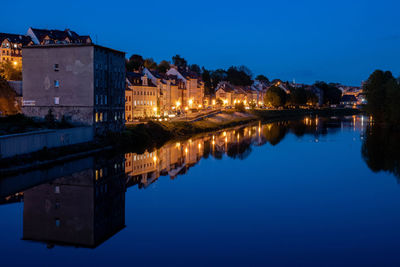 Illuminated buildings by river against sky at dusk