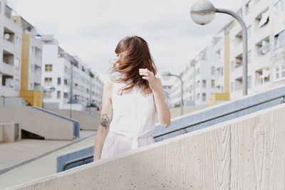 Young woman shaking head while standing by railing on steps against sky in city