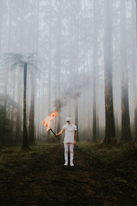 Full length of young man with fire standing in forest during foggy weather