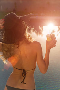 Rear view of shirtless young woman standing at beach during sunset