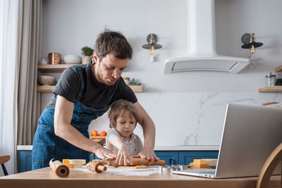 Cute preschool boy and his father baking cookies using video lesson on laptop